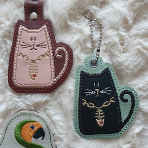 May include: Two embroidered cat keychains with a gold chain and a red jewel. The cat on the left is pink with brown accents and the cat on the right is black with green accents. A small white leather patch with a green parrot and the text "Sunthaga Designs" is in the lower left corner.