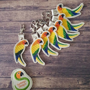 May include: A set of seven embroidered sun conure bird keychains with white leather backing and silver key rings. The birds are yellow, orange, green, and blue. The keychains are arranged in a semi-circle on a wooden surface. The image also includes a small, round, embroidered patch with the text "Sunthaga Designs".