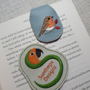 Op de afbeelding: Twee geborduurde leren bladwijzers. Eén is lichtblauw met een vogel en rode bessen. De andere is wit met een papegaaienontwerp en de tekst "Sunthega Designs" in oranje en groen.