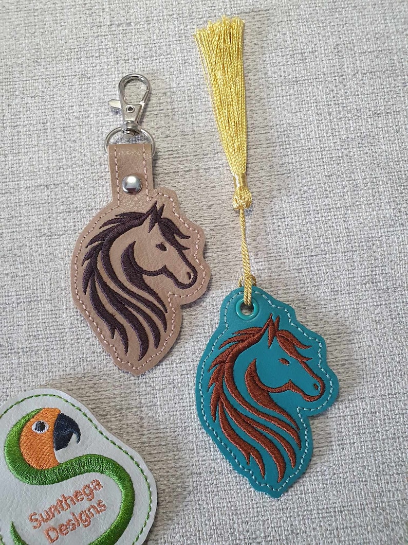 May include: Two leather keychains with horse head designs. One keychain is brown with a dark brown horse head and a silver key ring. The other keychain is teal with a brown horse head and a gold tassel. The keychains are on a light gray textured surface.