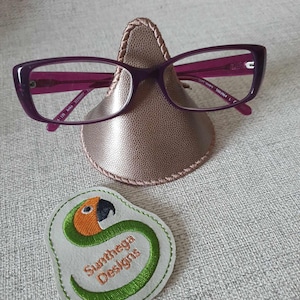 May include: A pink and gold glasses holder with a stitched edge. The holder is shaped like a nose and is holding a pair of purple glasses. A small white patch with a green snake and the text "Sunthera Designs" is in the lower left corner of the image.