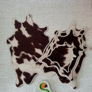 May include: Three brown and white cowhide coasters with a white background and brown spots. The coasters are stitched together with a white thread. A small green and orange logo with the text "Sunthaga Designs" is in the lower right corner of the image.