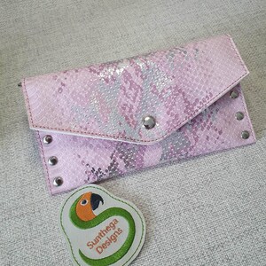 May include: Pink clutch with a snakeskin pattern and silver hardware. The clutch has a flap closure with a snap and is embellished with silver rivets. A small, embroidered patch with a parrot design and the text "Sunthega Designs" is also visible.