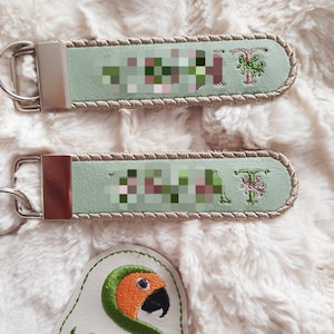 May include: Two light green leather keychains with a brown stitched border. Each keychain has a silver key ring and a floral design with the letter T embroidered in green. The bottom keychain has the word "Sunthaga Designs" embroidered in green on a white leather patch.