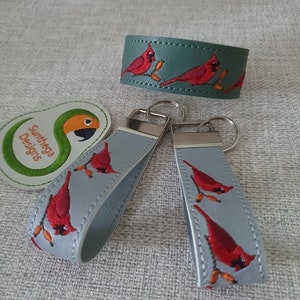 May include: A collection of accessories featuring embroidered cardinal designs. Includes two gray keychains, a green bracelet, and a patch with a parrot logo. The keychains and bracelet have red cardinal bird embroidery.