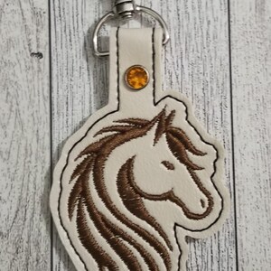 May include: A white leather keychain with a brown embroidered horse head design and a silver key ring. A small orange rhinestone is attached to the leather strap.