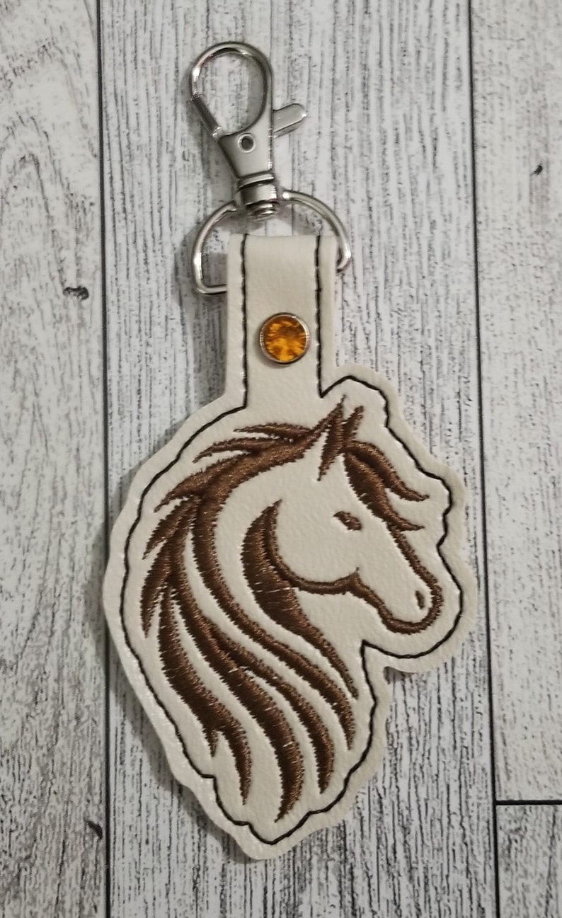 May include: A white leather keychain with a brown embroidered horse head design and a silver key ring. A small orange rhinestone is attached to the leather strap.