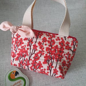 Könnte beinhalten: Kleine, gesteppte Tragetasche mit einem roten und braunen Blumenmuster auf cremefarbenem Hintergrund. Die Tasche hat einen roten Reißverschluss, cremefarbene Griffe und eine rosa Schleife. Ein gesticktes Patch mit einem Papageien-Design ist ebenfalls sichtbar.