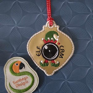 以下が含まれることがあります： 緑色の帽子をかぶってカメラを持ったエルフが描かれた、金色のクリスマスオーナメント。オーナメントには「Elf Cam」の文字が書かれている。オーナメントは白のドット模様の赤いリボンで吊り下げられている。小さな緑とオレンジの刺繍パッチには「Sunthaga Designs」の文字が書かれている。