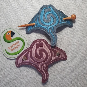 May include: Two embroidered hair barrettes in the shape of manta rays. One is blue with a wooden stick, the other is burgundy with star accents. A green and white patch with a parrot graphic is also visible.