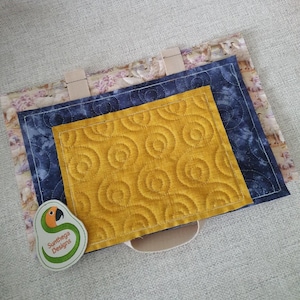 Puede incluir: Un bolso tote acolchado con un exterior estampado floral, un interior azul marino y un bolsillo acolchado amarillo mostaza. El bolsillo amarillo mostaza tiene un diseño en espiral. El bolso tiene asas beige y una etiqueta con un logotipo de loro y el texto "Sunthega Designs."