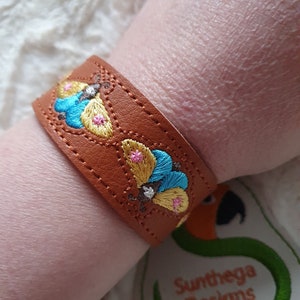 Könnte beinhalten: Ein braunes Lederarmband mit gestickten Schmetterlingen in Blautönen, Gelb und Rosa. Das Armband ist mit einem kleinen, gestickten Schmetterlingsdesign auf dem Leder verziert. Das Armband wird an einem Handgelenk gezeigt.