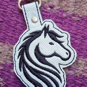 May include: White leather keychain with a black embroidered horse head design. The keychain has a silver metal clasp.