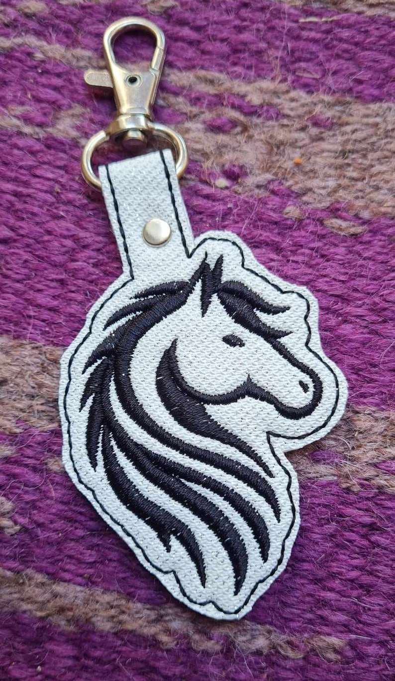 May include: White leather keychain with a black embroidered horse head design. The keychain has a silver metal clasp.