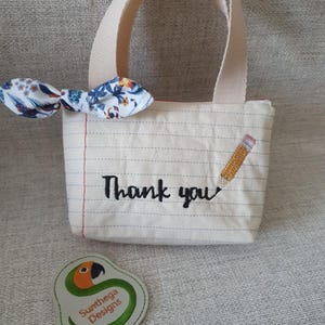 Könnte beinhalten: Eine kleine, cremefarbene Tragetasche mit einem linierten Papierdesign, mit den Worten "Thank you" und einer Bleistiftillustration. Eine florale Schleife ist an der Tasche befestigt. Ein Logo-Patch mit einem grünen Schlangenmuster ist ebenfalls vorhanden.