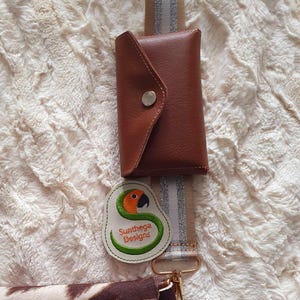 Puede incluir: Una cartera de cuero marrón con cierre de solapa y un botón plateado. La cartera está unida a una correa a rayas beige y plateada. Un parche bordado verde y naranja con el texto "Sunthaga Designs" está unido a la correa.