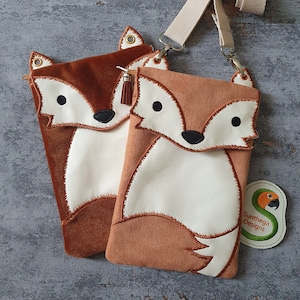 May include: Two fox-shaped crossbody bags with a brown and white design. The bag on the left is made of brown velvet and the bag on the right is made of brown fabric with a herringbone pattern. Both bags have a white fox face with black eyes and a black nose. The bag on the right has a white tail with a brown tip. The bags are attached to a white strap. The bag on the right has a green tag with the text "Sunthaga Designs" and a parrot illustration.