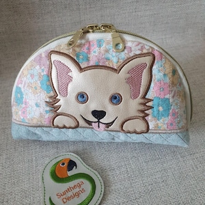 May include: A white zippered pouch with a floral pattern and a white puppy peeking out. The pouch has a gold zipper and a green embroidered patch with the text "Sunthaga Designs" and a parrot.