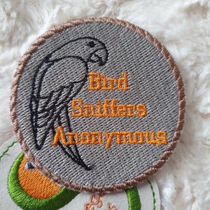 May include: A gray embroidered patch with a brown border. The patch features a black silhouette of a bird with the text "Bird Stuff Anonymous" in orange embroidery.