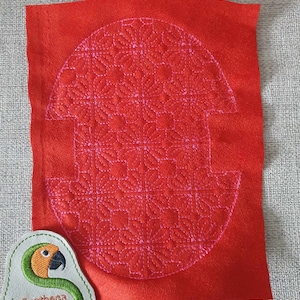 Könnte beinhalten: Ein quadratisches Stück roter Filzstoff mit einem rosa gestickten Muster aus geometrischen Formen. Das Muster hat die Form eines abgerundeten Rechtecks mit abgerundeter Oberseite und gerader Unterseite.