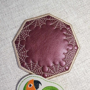 May include: A burgundy leather coaster with a decorative stitched design in a light brown thread. The coaster is shaped like an octagon with rounded edges.
