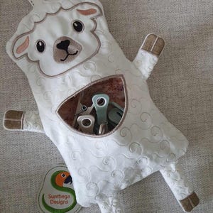 May include: White plush lamb-shaped toy with a pocket for storing small items. The toy has a brown embroidered face and a loop for hanging. The text "Sunthaega Designs" is on a green and orange parrot-shaped tag.