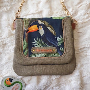 Peut inclure: Un sac à bandoulière en similicuir gris avec un imprimé toucan et une bandoulière en chaîne dorée. Le sac a un rabat avec une bordure en cuir marron et une petite étiquette qui dit "Handmade".