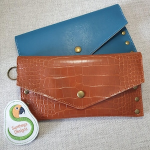 May include: Two leather wallets in teal and brown. The brown wallet has a crocodile texture and a brass snap closure. The teal wallet is smooth with brass studs. A white and green tag with a parrot design and the text "Sunthega Designs" is also visible.