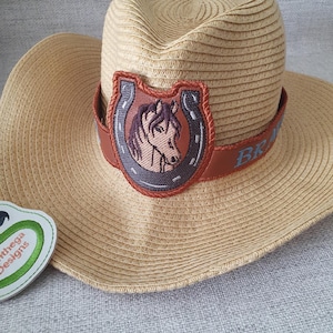 Könnte beinhalten: Ein Strohhut im Cowboy-Stil mit einem braunen Lederband und einem Hufeisen mit einem Pferdekopf-Design. Das Band ist mit dem Wort "BR" bestickt.