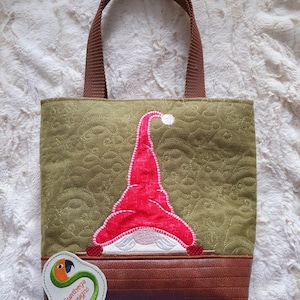 May include: A green quilted tote bag with a red gnome design. The gnome has a white beard and a red hat with a white pom-pom. The bag has brown handles and a brown leather-like bottom. A tag with the text "Sunthega Designs" is attached.