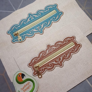 May include: Two decorative zippered pouches, one teal and one brown, with ornate scrollwork embroidery. Each pouch has a gold-coloured zipper. A small, avocado-shaped patch with a parrot design and the text "Sunthega Designs" is also visible.