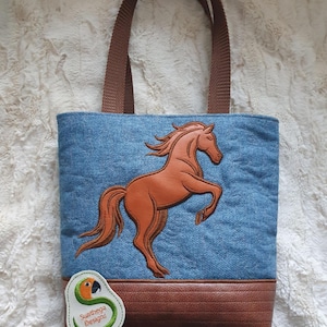 May include: A denim tote bag with a brown leather horse applique. The bag has brown handles and a brown leather bottom. A small tag with a parrot logo and the text "Sunthega Designs" is attached.
