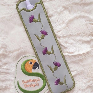 Può includere: Un portapenne in tessuto grigio con un bordo ricamato verde e fiori di cardo viola. Il portapenne è appoggiato su una superficie bianca e pelosa. Una toppa ricamata verde con un pappagallo e il testo "Sunthaga Designs" si trova nell'angolo in basso a sinistra dell'immagine.