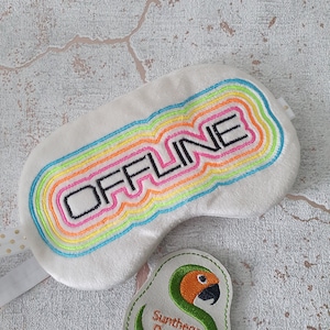 Könnte beinhalten: Weiße Schlafmaske mit dem Wort "OFFLINE" in Schwarz mit Regenbogenfarben umrandet gestickt. Ein grünes gesticktes Patch mit einem Papagei und dem Text "Synthega Designs" befindet sich in der unteren rechten Ecke.