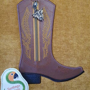 May include: A brown cowboy boot-shaped pouch featuring a vertical zipper closure with a silver-toned dog head charm. The boot is decorated with yellow stitched patterns and has a black heel. A small white patch with a parrot logo and the text Sunthega Designs.