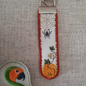 May include: White leather keychain with a red border. The keychain features an embroidered design of a spider on a web, a pumpkin, and a vine with leaves and flowers. The keychain has a silver key ring.