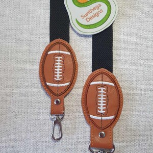 Könnte beinhalten: Zwei braune Leder-Schlüsselanhänger mit einem weißen gestickten Fußball-Design. Jeder Schlüsselanhänger hat einen silbernen Metallclip.
