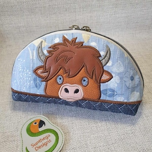 Puede incluir: Una bolsa de cremallera de tela azul y blanca con un rostro de vaca Highland bordado en marrón y blanco. La bolsa está decorada con flores y hojas blancas. Un pequeño parche de cuero marrón con el texto "Sunthaya Designs" y un loro se encuentra en la esquina inferior izquierda de la imagen.