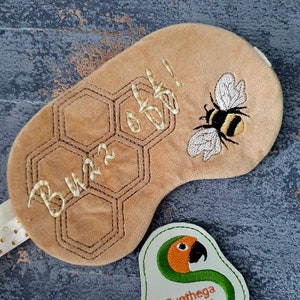 Puede incluir: Una máscara de dormir marrón con un patrón de panal y el texto "Buzz off!" bordado en hilo dorado. Una abeja está bordada en el lado derecho de la máscara. Una etiqueta blanca con un loro verde y el texto "Sunthega Designs" está en la esquina inferior derecha de la imagen.