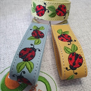 May include: Three leather wristbands and keychains with ladybug embroidery. The wristband is light yellow, and the keychains are light blue and yellow. Each features a red ladybug with black spots and green leaves.