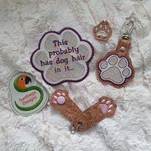 May include: A set of five embroidered paw print patches. The patches are brown with white paw prints and pink accents. One patch has the text "This probably has dog hair in it..." in purple lettering. The patches are on a white fluffy surface.