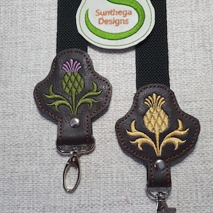 May include: Two brown leather keyrings featuring thistle designs, one with purple and green detailing, the other with gold. They have silver clasps and are shown with a black strap and a logo with a parrot.