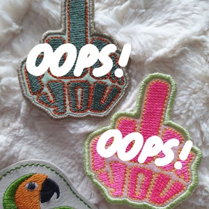 Puede incluir: Parches bordados con forma de mano haciendo la peineta, con el texto "oops!" y "F*ck You". Un parche es verde y naranja, el otro rosa y amarillo. También se ve un tercer parche con un loro.