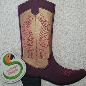 Könnte beinhalten: Ein dekorativer Aufnäher in Form eines Cowboystiefels mit dunkelvioletter Basis, metallisch goldenem Schaft und pinkfarbenen Stickereidetails. Daneben befindet sich ein runder Aufnäher mit einem grünen Papageienlogo und dem Text Sunthega Designs.