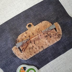 Op de afbeelding: Een bril ligt in een etui met kurkpatroon en een knoopsluiting. De bril heeft een zilverkleurig montuur met decoratieve accenten. Een kleine patch met de tekst "Sunthega Designs" en een papegaaienontwerp is ook zichtbaar.