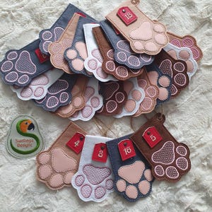 Peut inclure: Un ensemble de 18 décorations en feutre en forme de patte de chien avec des numéros rouges sur des étiquettes. Les pattes sont de différentes couleurs, notamment le brun, le gris, le blanc et le rose. Les décorations sont enfilées ensemble avec de la ficelle.