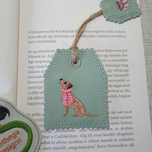 Könnte beinhalten: Hellgrüner Leder-Lesezeichen mit genähter Bordüre, das einen gestickten braunen Hund mit rosa Schal zeigt. Das Lesezeichen ist mit einer Schnur an einem Buch befestigt. Ein kleineres Etikett mit einem Pfotenabdruck ist ebenfalls angebracht.