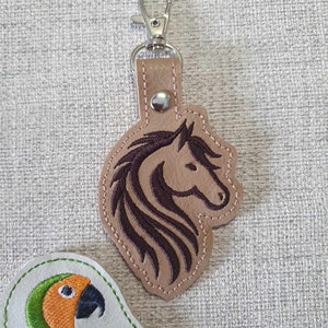 May include: A brown leather keychain with a dark brown embroidered horse head design. The keychain has a silver metal clasp.