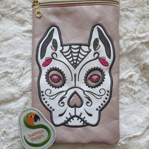 May include: A light pink crossbody bag with a zipper closure. The front of the bag features an embroidered design of a white sugar skull with black and pink details. A small white patch with a green parrot logo and the text "Sunthaga Designs" is attached to the bottom of the bag.