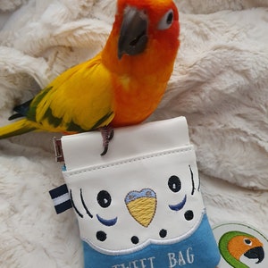 以下が含まれることがあります： 青い鳥の形をしたポーチで、前面に「TWEET BAG」という文字が刺繍されています。ポーチの上に、太陽コンゴウインコがとまっており、カメラを見ています。